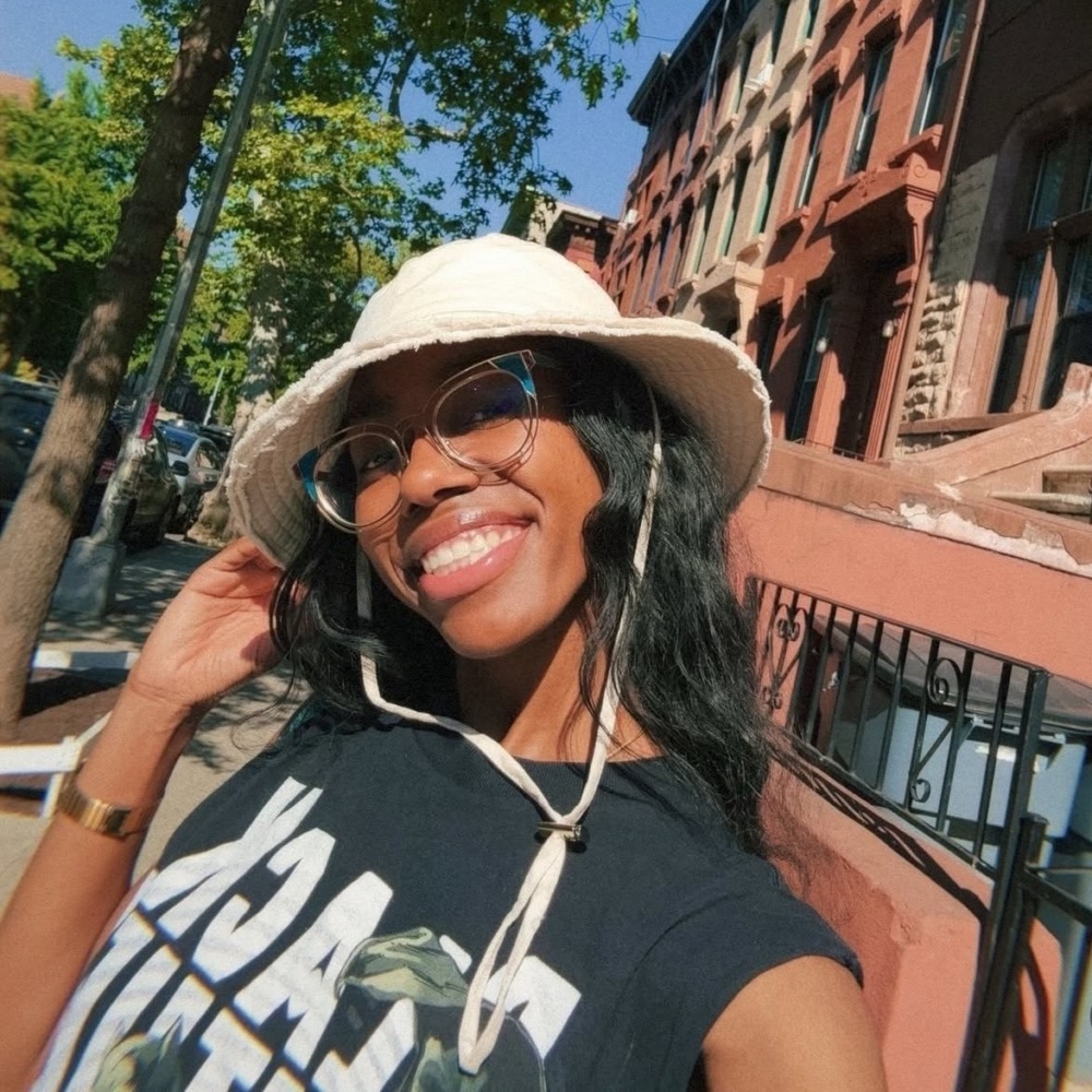A person wearing glasses and a bucket hat smiles while taking a selfie outdoors on a sunny day in a residential neighborhood.