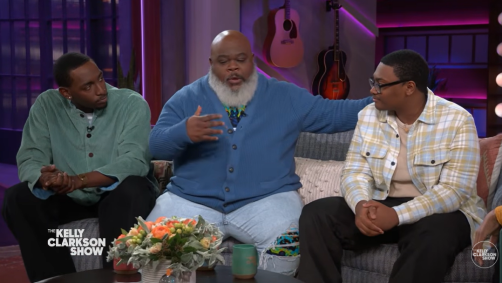 Three men sit on a couch having a discussion on The Kelly Clarkson Show set, with two guitars and flowers visible in the background.
