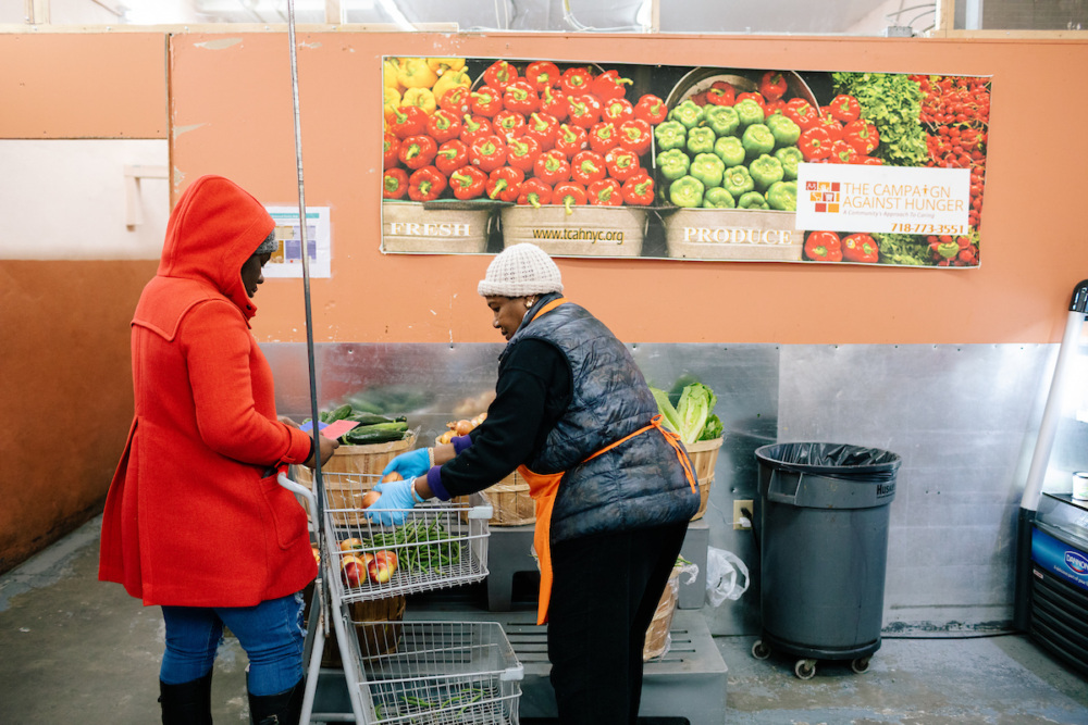 Two women stand by produce bins in a food pantry; one hands groceries to the other. A sign above displays fresh vegetables and reads "The Campaign Against Hunger.