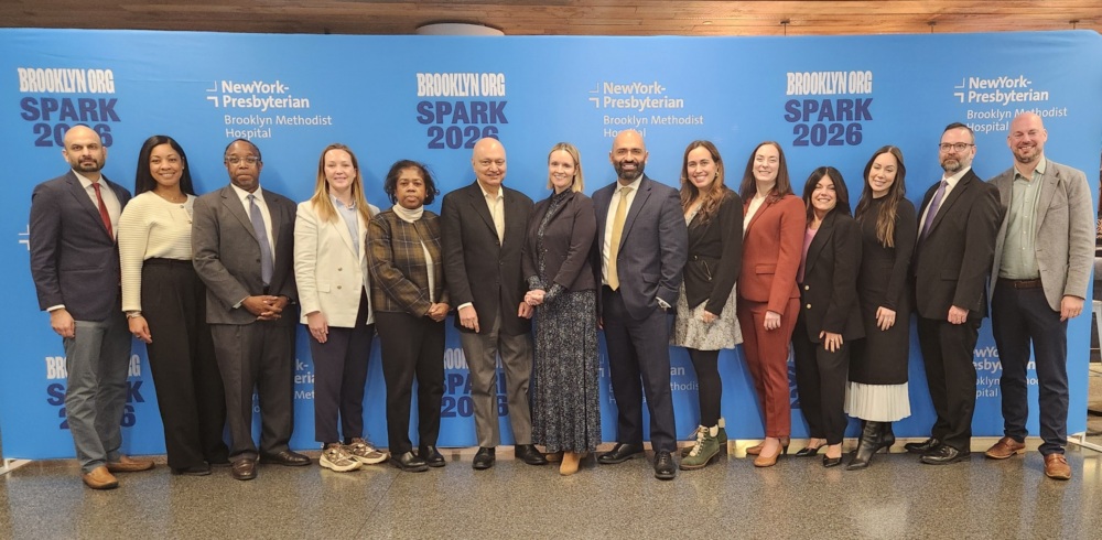 Thirteen people stand in a line posing for a group photo in front of a blue "SPARK 2026" and "NewYork-Presbyterian Brooklyn Methodist Hospital" backdrop.
