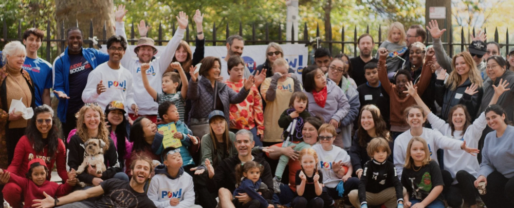 A large group of adults and children pose together outdoors in front of a fence, some holding signs and wearing shirts with a "POW!" logo.