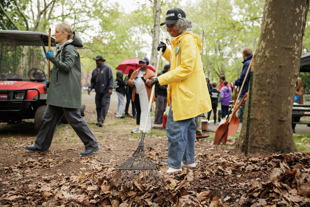 A group of people rake fallen leaves in a park during an outdoor community clean-up event.