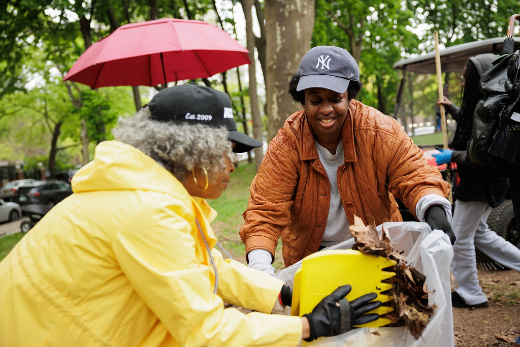 Two people wearing gloves collect dry leaves in a yellow bucket and place them into a trash bag during a park cleanup on a cloudy day.