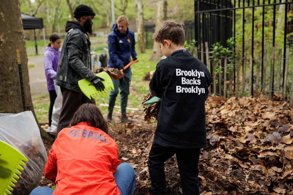 A group of people, including children and adults, gather leaves and debris in a park, wearing jackets and gloves, participating in a community cleanup.