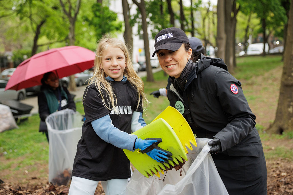 Two people, one child and one adult, wearing gloves and black shirts, collect leaves in a park using a yellow plastic scoop. Other volunteers and trees are visible in the background.