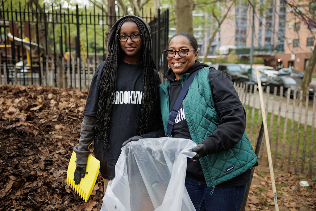 Two women wearing gloves and "Brooklyn" shirts collect leaves in a plastic bag in an outdoor park area with a fence and trees in the background.