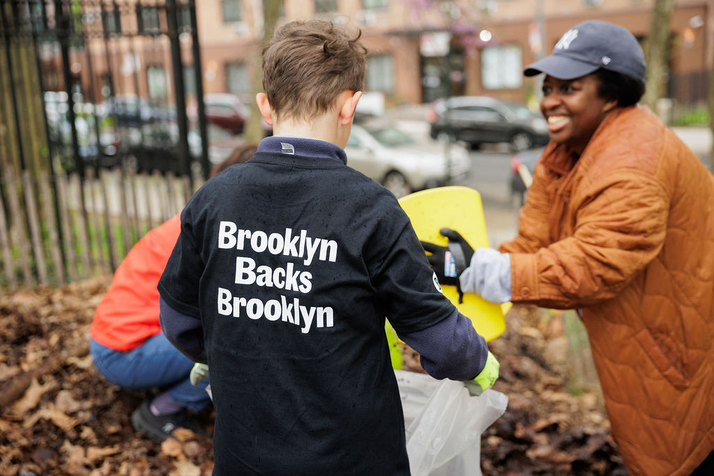 A person wearing a "Brooklyn Backs Brooklyn" shirt gathers leaves with others in an outdoor community cleanup.