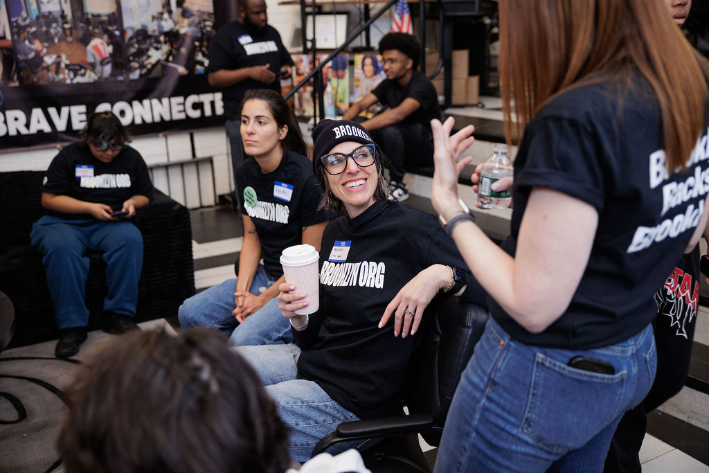A group of people wearing matching "BROOKLYN.ORG" shirts sit and talk in a casual indoor setting; one person smiles while holding a coffee cup.