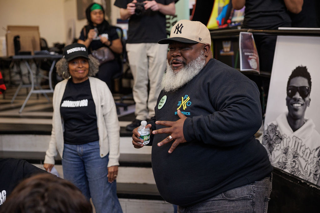 A man in a black sweatshirt and beige cap speaks to a group indoors, holding a water bottle. A woman stands nearby, and a black-and-white portrait photo is visible in the background.
