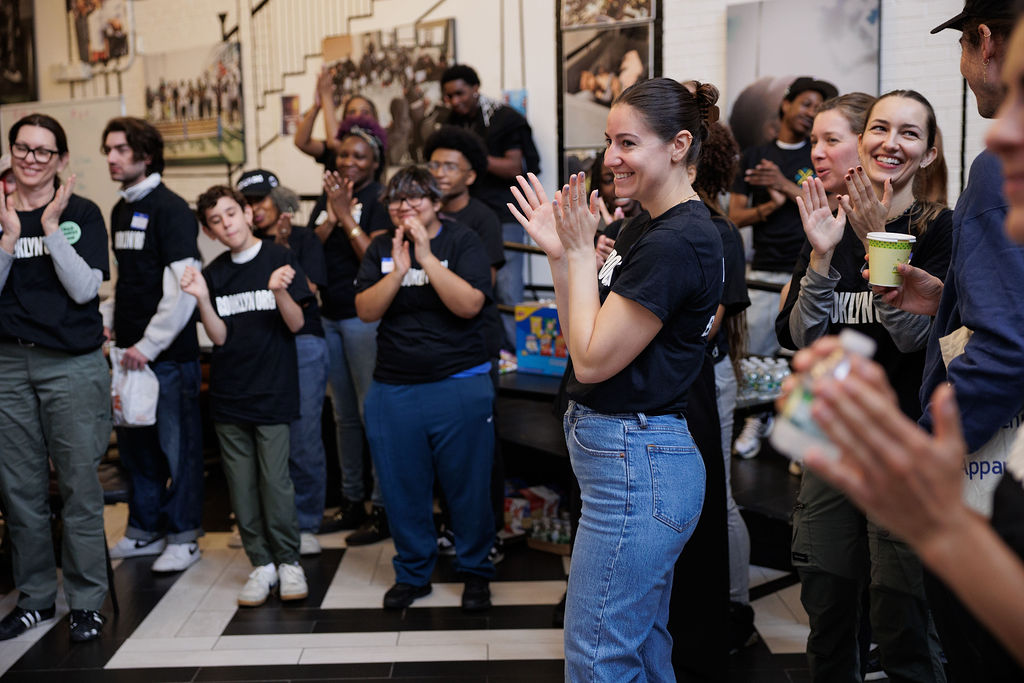 A group of people in casual clothes stand indoors, clapping and smiling, appearing to participate in a group event or gathering.