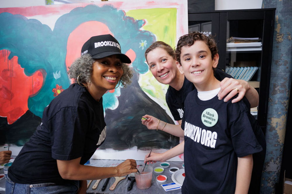 Three people wearing matching "BROOKLYN.ORG" shirts paint together in front of a colorful mural, smiling at the camera. Art supplies are visible on the table.