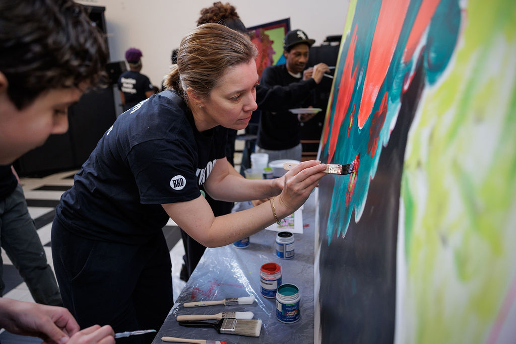 A woman paints a colorful mural on a large canvas, surrounded by other people working on art projects in a studio setting.
