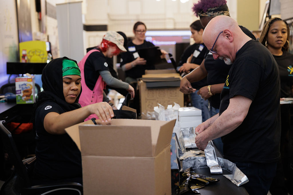 A group of people wearing black shirts assemble care packages at tables, placing items into boxes and organizing supplies.