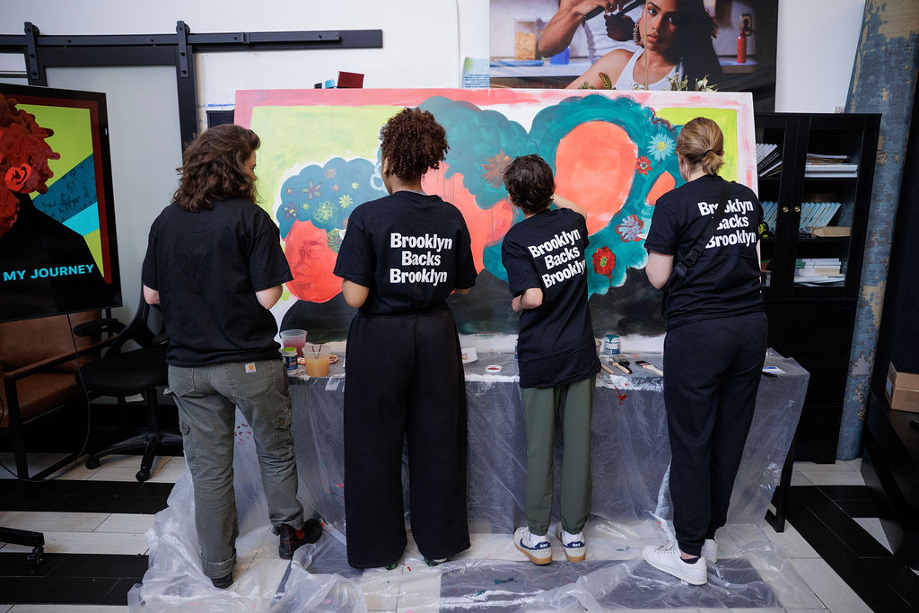 Four people with “Brooklyn Backs Brooklyn” shirts paint together on a large, colorful mural indoors, standing with their backs to the camera.
