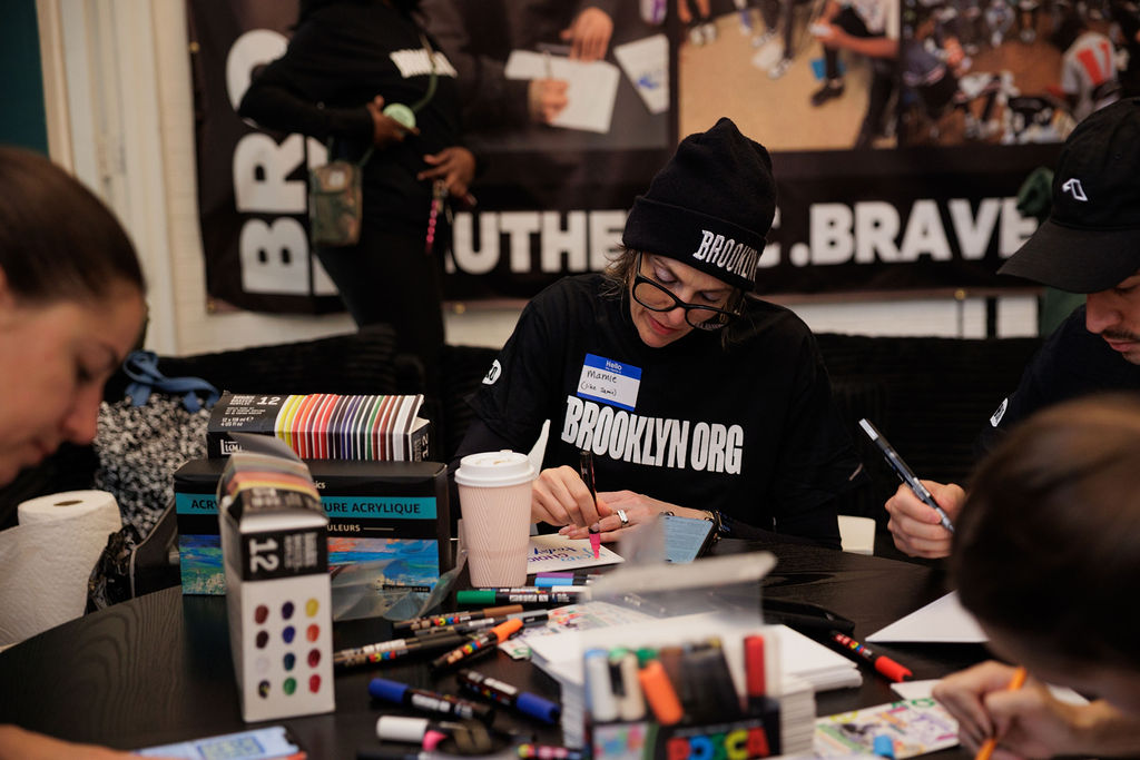 People sit around a table drawing and coloring with markers and colored pencils. A person wearing a "BROOKLYN.ORG" shirt and beanie is focused on their artwork. Art supplies are scattered on the table.