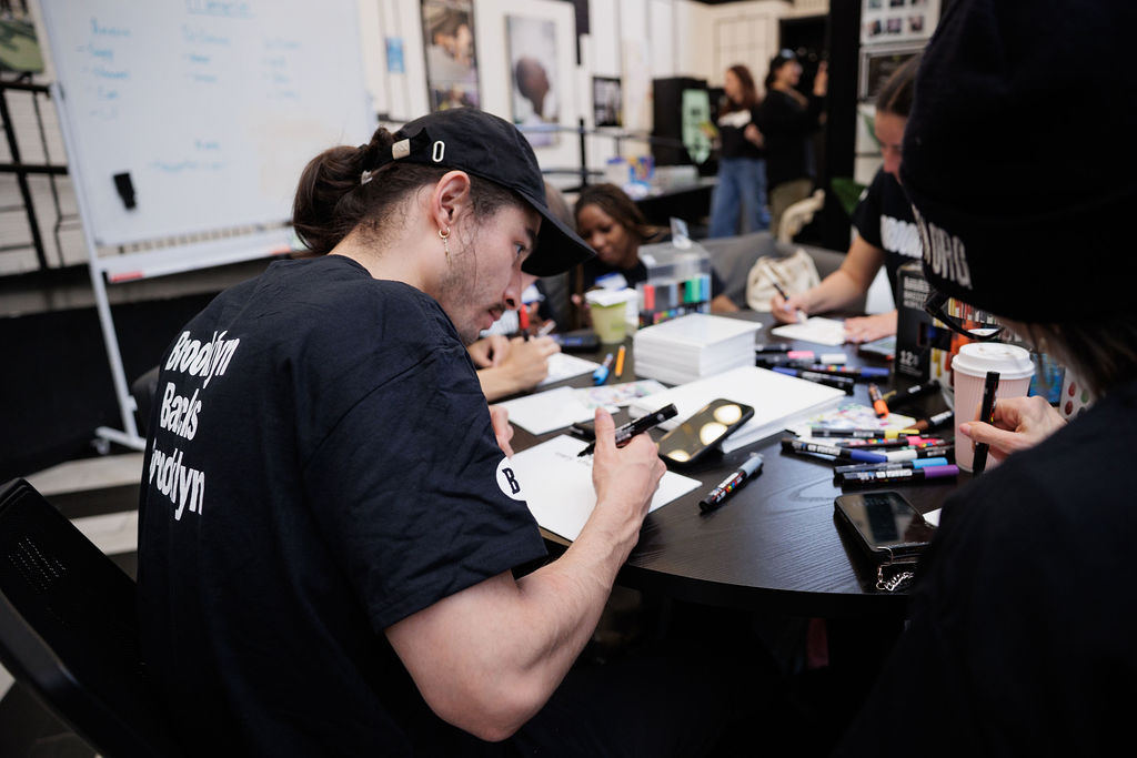 People sit around a table working with markers and paper, with art supplies and coffee cups visible; a whiteboard and photos are in the background.