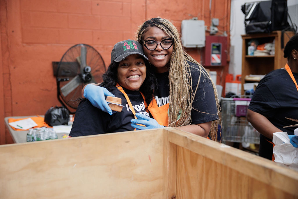 Two women wearing gloves and aprons smile and hug in a warehouse setting, standing behind a large wooden crate.