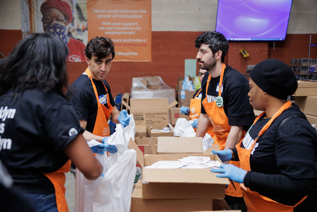 Four volunteers wearing gloves and orange aprons pack food items into bags at an indoor food bank. Boxes and supplies are visible on the tables and shelves around them.