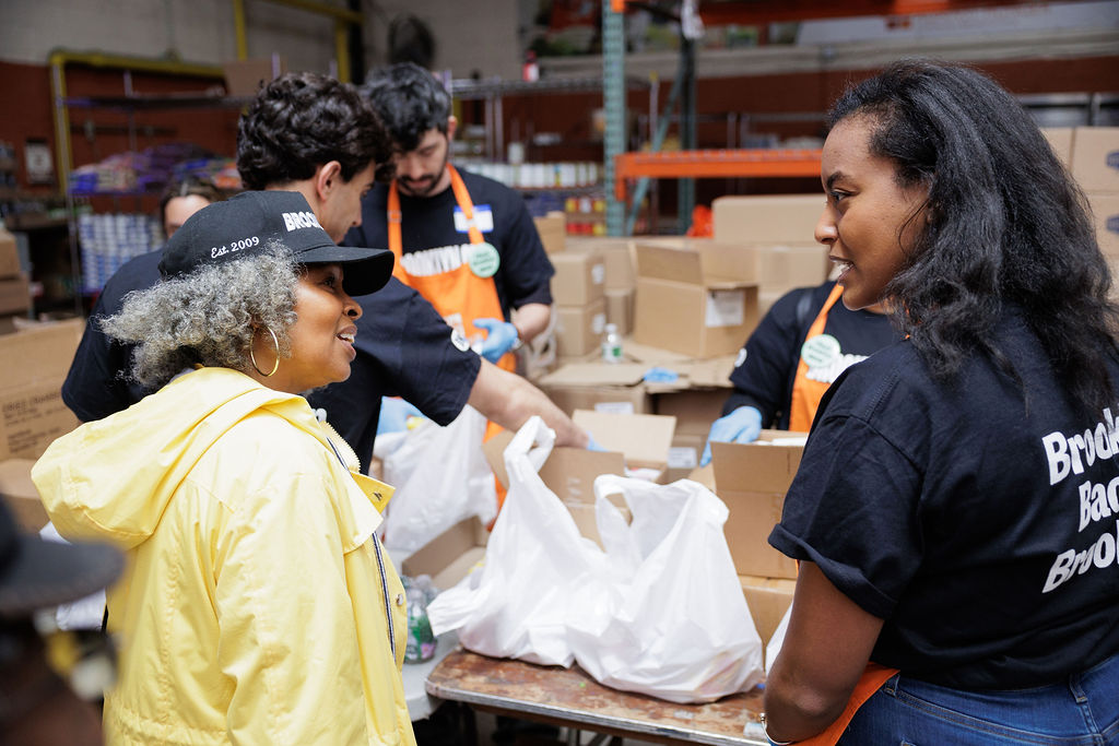 Volunteers in a warehouse pack supplies into bags while two women in the foreground have a conversation. Cardboard boxes and bags are on the table.