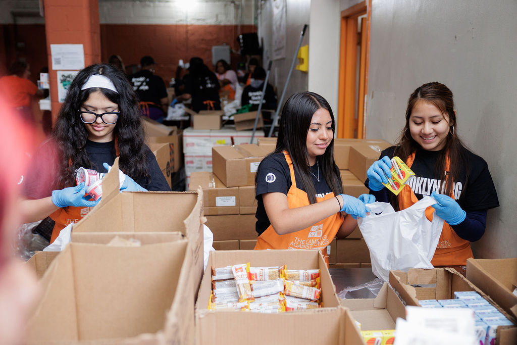 Three volunteers wearing gloves pack food items into bags at a distribution center, surrounded by boxes of supplies.