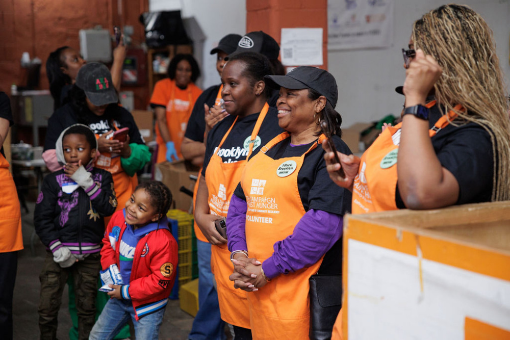 A group of volunteers wearing orange aprons stand together and smile in a warehouse, with children present and boxes in the background.