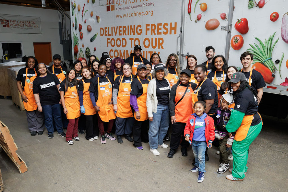 A diverse group of volunteers wearing orange aprons poses together in front of a food delivery truck with a "Delivering Fresh Food to Our Community" sign.