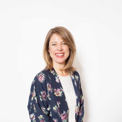 Woman with shoulder-length blonde hair, wearing a floral-patterned navy top and white blouse, smiling in front of a plain white background.
