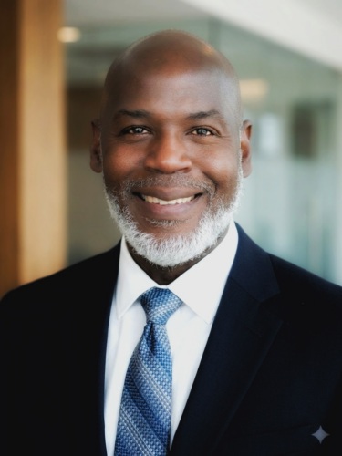 A man in a suit and tie smiles at the camera in an indoor office setting with blurred background.