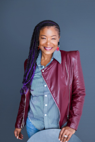 A woman with long braided hair, wearing a maroon leather jacket over a striped shirt and blue jeans, stands smiling in front of a plain gray background.