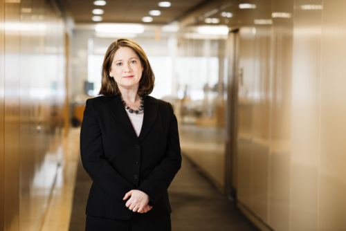 A woman in a black suit stands in a well-lit hallway with glass walls, looking at the camera with a neutral expression.