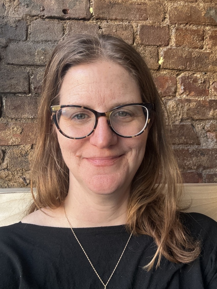 A woman with glasses and long brown hair, wearing a black top and a necklace, sits in front of a brick wall and smiles slightly at the camera.