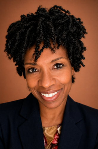 A woman with curly hair and a dark blazer smiles at the camera against a plain brown background.