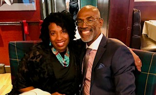 A man in a suit and a woman in a black top with turquoise jewelry sit together and smile at a restaurant booth.