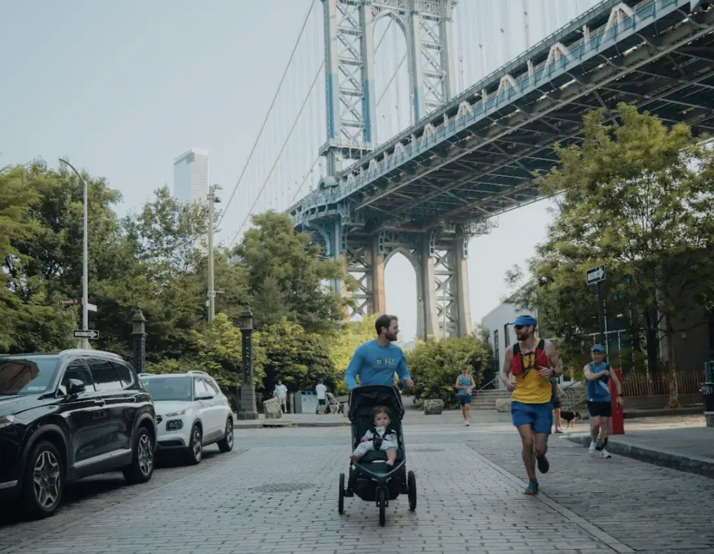A man pushes a child in a stroller on a cobblestone street as two runners pass by, with a large bridge and trees in the background.