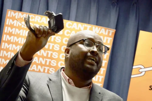 A man in glasses and a suit holds up a broken restraint device, speaking in front of signs that read "IMMIGRANTS CAN'T WAIT.