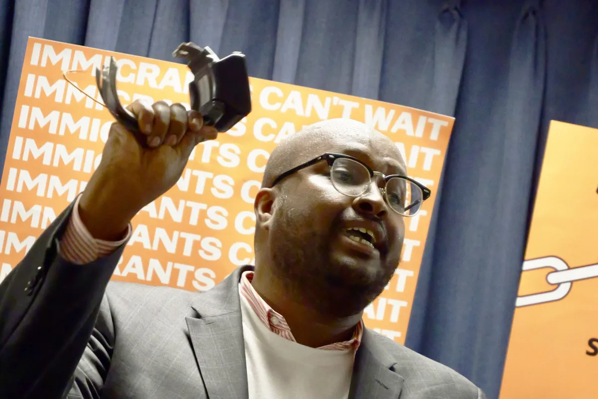 A man in glasses and a suit holds up a broken restraint device, speaking in front of signs that read "IMMIGRANTS CAN'T WAIT.
