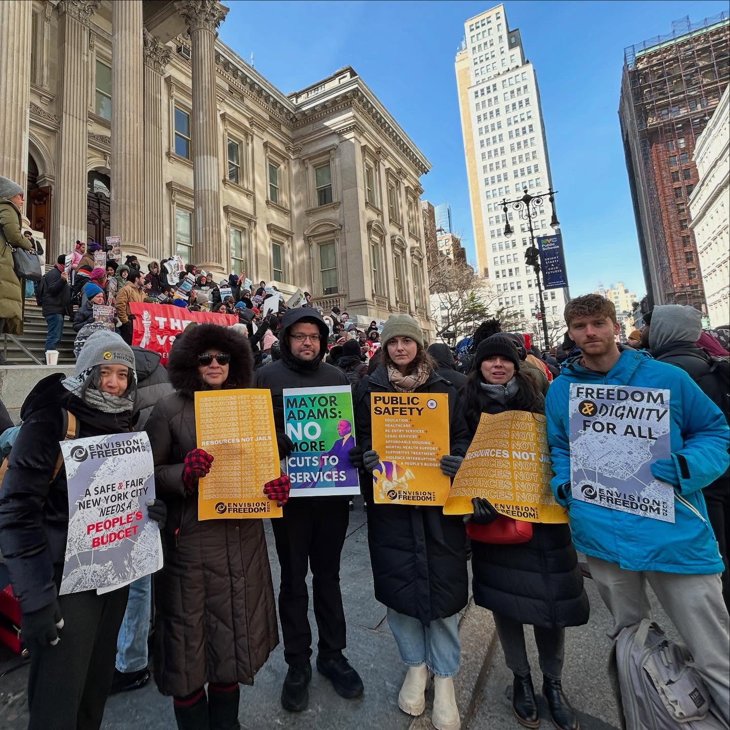 A group of six people stand outside a building holding protest signs about service cuts, public safety, and freedom and dignity for all. More people and banners are visible on the steps behind them.