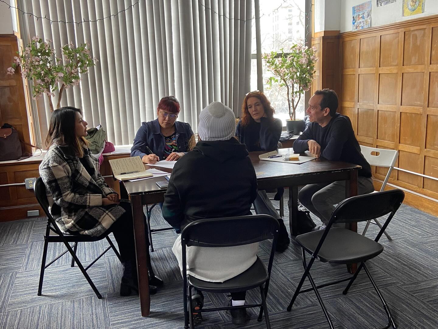 Five people sit around a rectangular table in an office, engaged in discussion. Papers, notebooks, and a coffee cup are on the table.