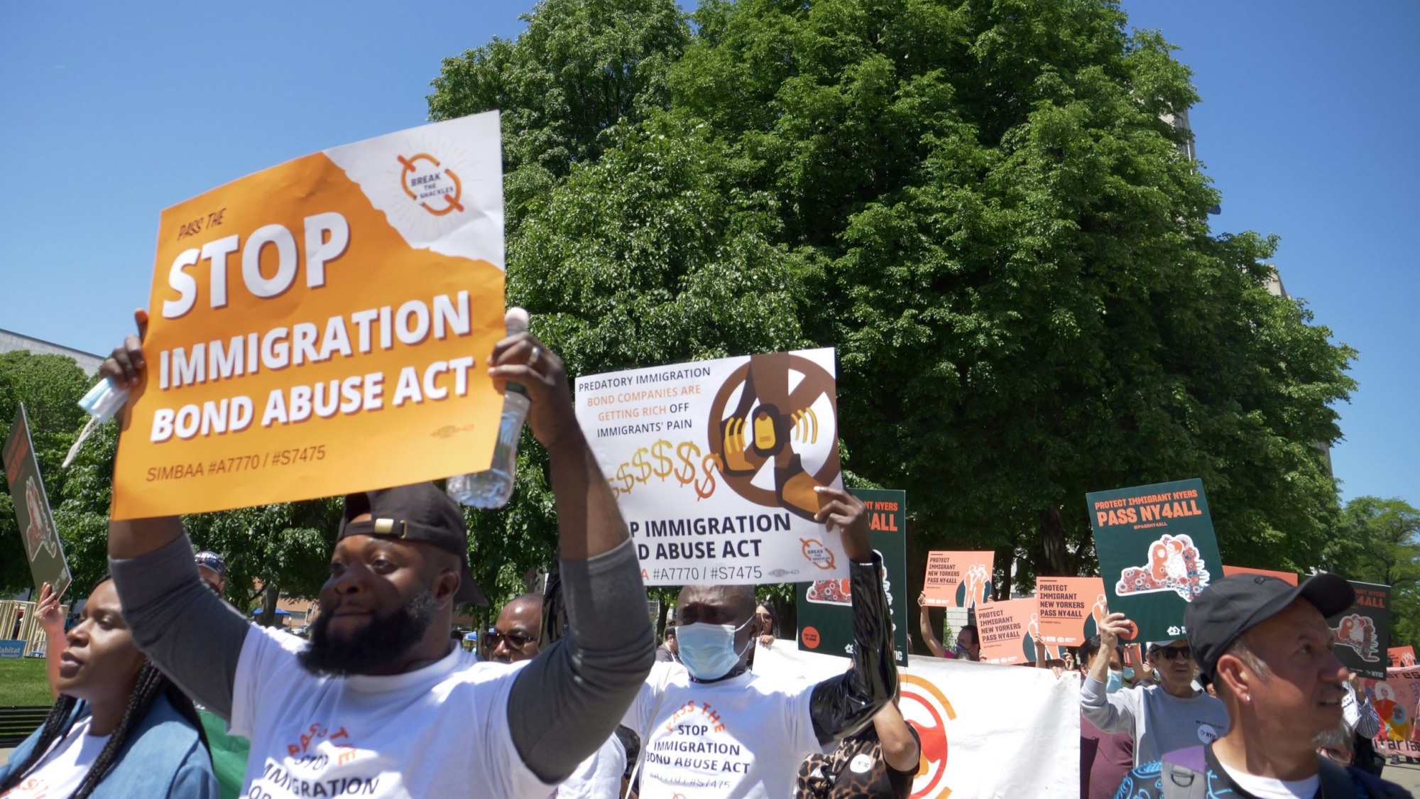 A group of protesters hold signs opposing the Immigration Bond Abuse Act during a daytime rally outdoors, with trees visible in the background.