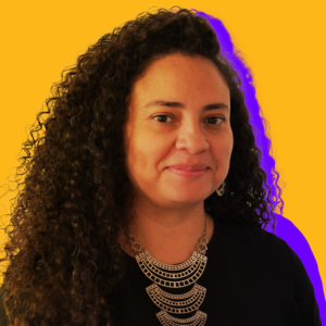 A woman with curly hair wearing a black top and a large metallic necklace stands against a vibrant yellow and purple background.