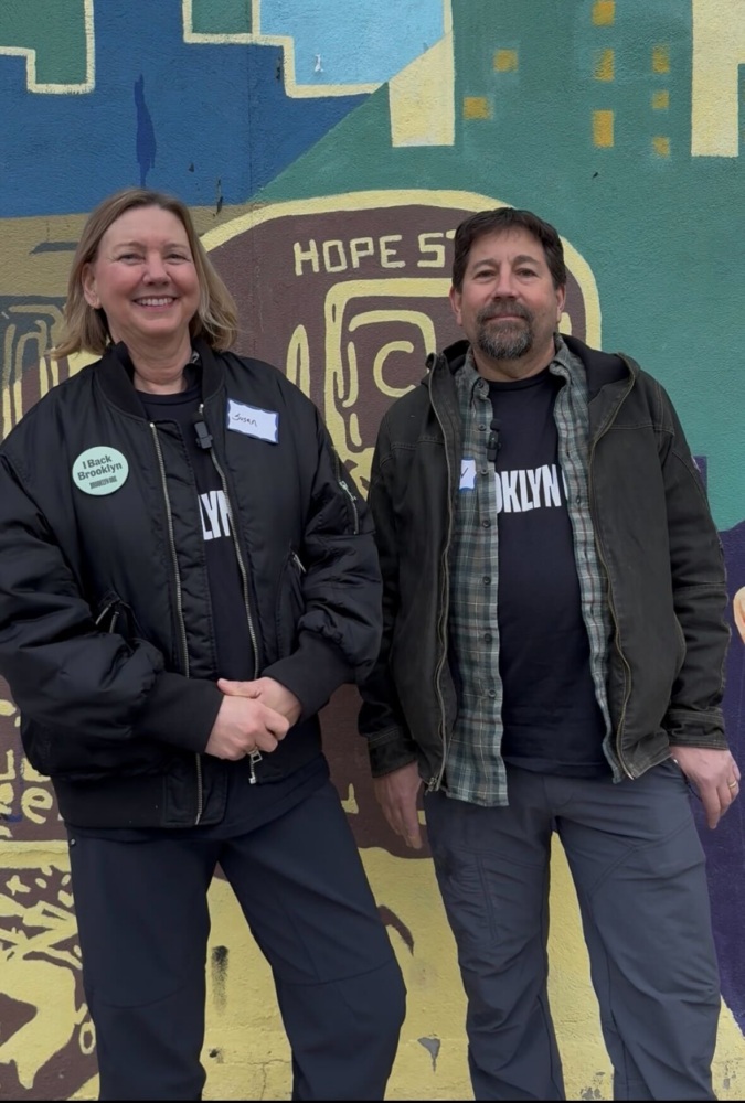 Two people standing side by side in front of a colorful mural, both wearing dark jackets and name tags, one with a sticker that says "I Block Brooklyn.