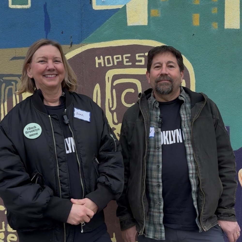 Two people stand in front of a colorful mural. Both wear dark jackets and "Brooklyn" t-shirts; one has a "I block the BQE" button. They both have name tags and are smiling slightly.