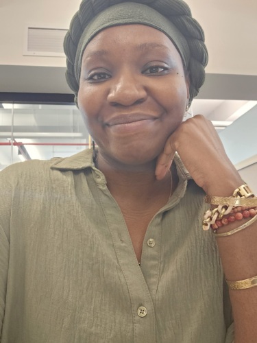 A woman wearing a green headwrap and green shirt smiles at the camera, resting her chin on her hand. She wears bracelets and a watch on her wrist.