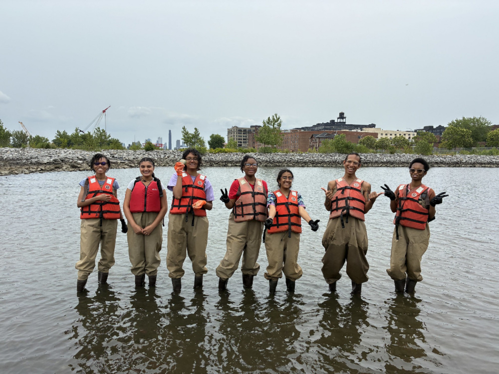 Seven people wearing orange life vests and waders stand in shallow water, posing for a group photo with buildings and trees visible in the background.