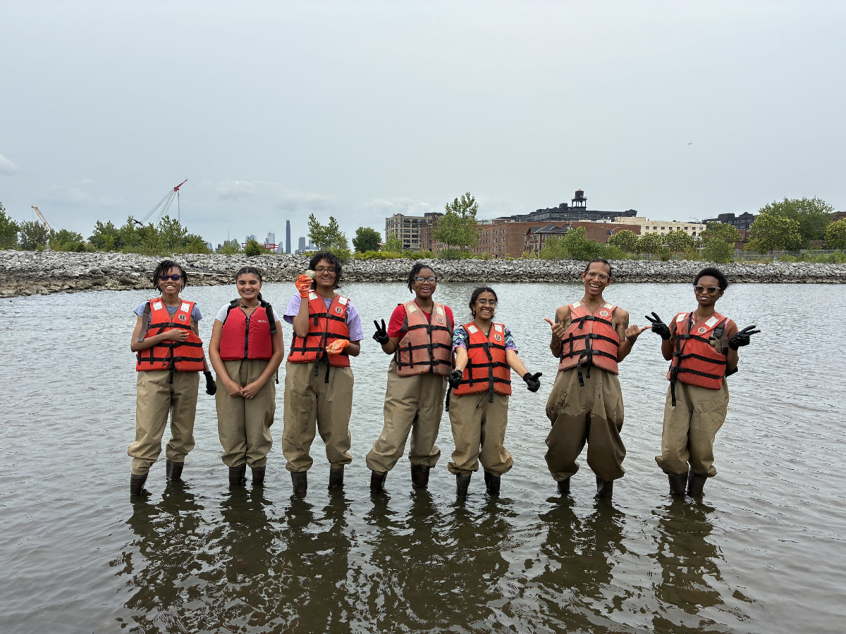 Seven people wearing orange life vests and waders stand in shallow water, posing for a group photo with buildings and trees visible in the background.