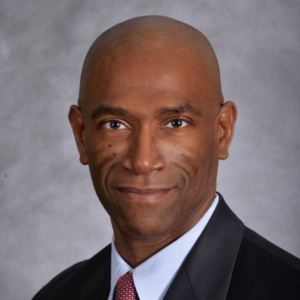 A man in a suit and red tie poses for a professional headshot against a gray background.