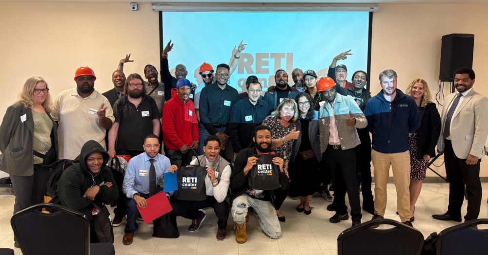 A diverse group of people pose together indoors in front of a projector screen displaying "RETI," some holding tote bags and wearing hard hats.