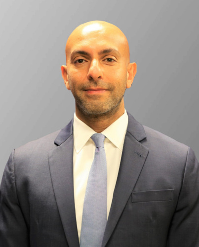 A man in a suit and tie stands against a plain grey background, looking at the camera with a neutral expression.