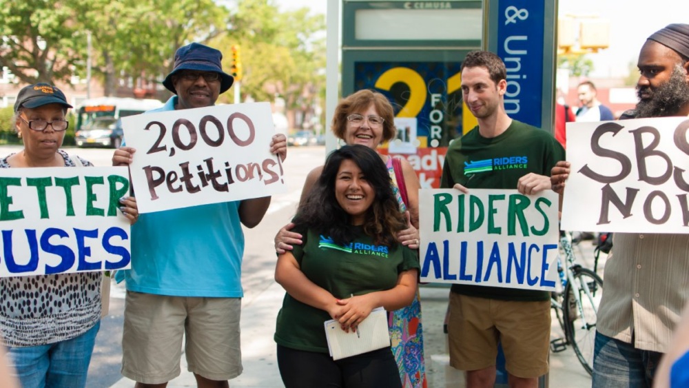 A group of people hold signs reading “2,000 Petitions!,” “RIDERS ALLIANCE,” and “SBS NOW!” at an outdoor event, advocating for better bus services.
