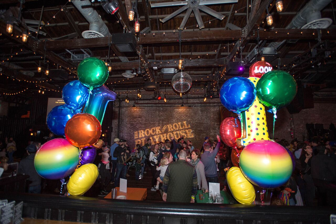 People gather at an indoor venue decorated with rainbow balloons, colorful lights, and a disco ball, with "Rock & Roll Playhouse" projected on a brick wall.
